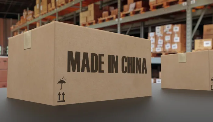 A close-up of a cardboard box with the text "MADE IN CHINA" and shipping symbols, set against a blurred warehouse background with more boxes.