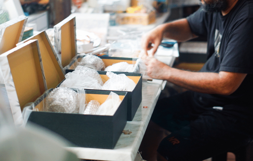 A worker in a warehouse carefully assembling and packing items into black boxes lined with protective wrapping at a kitting station.
