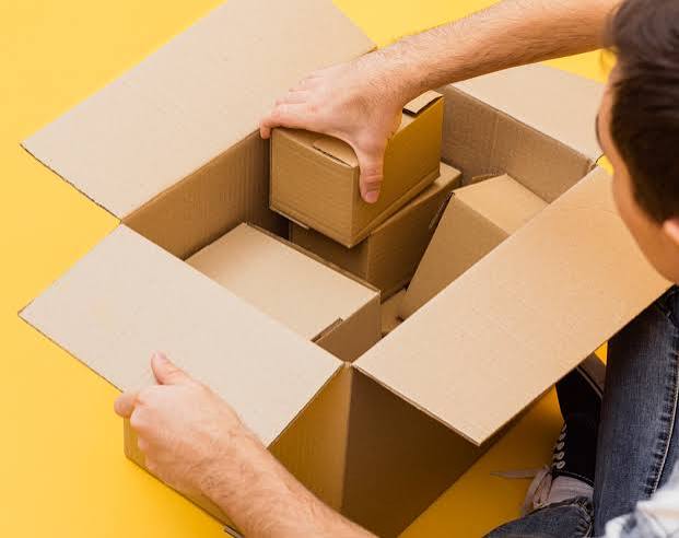 A person packing a small cardboard box into a larger open cardboard box on a bright yellow surface, illustrating kitting and bundling.